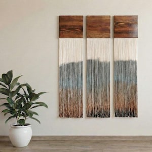 Op de afbeelding: Driedelige wandkunst met houten toppen en hangende strengen. De strengen gaan over van wit naar blauw, grijs en bruin. Het kunstwerk wordt tentoongesteld op een neutrale muur, met een plant in pot op de voorgrond. Moderne decoratie.