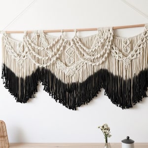 Puede incluir: Un tapiz de macramé con una combinación de colores natural y negro. La sección superior presenta intrincados patrones tejidos, mientras que la mitad inferior hace la transición a una franja negra. Se suspende de una barra de madera con un cordón blanco.