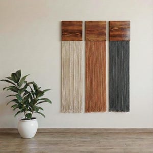 May include: Three wall hangings with wooden tops and textured fringe in cream, rust, and grey. The wall art is displayed on a neutral wall above a wooden floor. A potted plant with green leaves sits to the left.