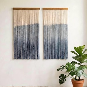 May include: Two wall hangings with a gradient design. The top portion is a light beige, transitioning to a deep blue at the bottom. Each piece is mounted on a wooden bar. A potted plant with large green leaves sits to the right.