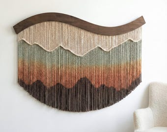 May include: A large, woven wall hanging with a dark brown, curved wooden top. The design features layers of yarn in shades of beige, green, orange, and brown, creating a mountain-like pattern. The bottom edge is a semi-circle.