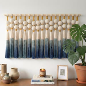 May include: A large wall hanging with a wooden dowel, featuring white, yellow, and blue tassels. The design includes large white spheres and a gradient effect on the tassels, transitioning from white to deep blue. A Monstera plant in a terracotta pot is to the right.