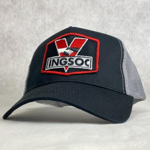 May include: Black and grey trucker hat with a red and white patch that says "INGSOC" and features a stylised "V" and a handshake.