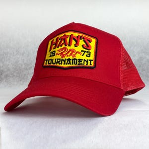 Könnte beinhalten: Rote Mesh-Trucker-Kappe mit einem gestickten gelben und roten Patch, auf dem "HAN'S 19 73 TOURNAMENT" mit einem roten Drachen in der Mitte steht.
