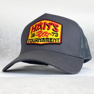 May include: A grey trucker cap with a mesh back and a curved peak. The front displays a rectangular patch with the text "HAN'S 1973 TOURNAMENT" in red and yellow, with a dragon graphic.