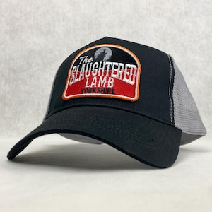 Peut inclure: Casquette de camionneur noire et grise avec un patch brodé rouge, blanc et orange qui dit "The Slaughtered Lamb Yorkshire".