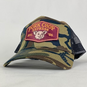 May include: Camouflage trucker hat with a black mesh back and a curved peak. The front has a rectangular patch with "Pork Chop Express" and a pig illustration in burgundy and gold. The hat is a classic accessory.