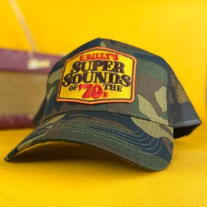 May include: A camouflage trucker hat with a black mesh back and a curved brim. The front features a patch with the text "K-Billy's Super Sounds of the 70's" in yellow, orange, and red. The hat is set against a yellow background.