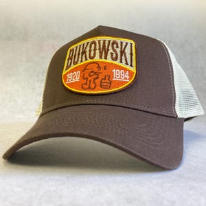 Puede incluir: Una gorra de camionero marrón y blanca con un parche marrón, naranja y amarillo que dice "Bukowski 1920 - 1994" con una imagen de dibujos animados de un hombre con un casco.