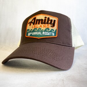 Op de afbeelding: Bruine en witte truckercap met een patch waarop "Amity Island 50th Annual Regatta" staat in oranje, groene en witte tekst.