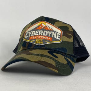Pode incluir: Boné de beisebol camuflado com parte traseira em malha preta. A frente apresenta um patch com o texto "CYBERDYNE SYSTEMS EST. 1984" e um gráfico de montanha em laranja, branco e cinza.