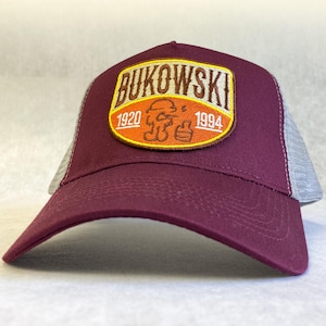 Könnte beinhalten: Eine burgunderrote und graue Trucker-Kappe mit einem braunen Patch mit dem Text "BUKOWSKI 1920 1994" und einer Cartoon-Illustration einer Person.