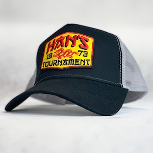 May include: Black and grey trucker hat with a yellow and red embroidered patch that says "HAN'S 19 73 TOURNAMENT" with a red dragon design.