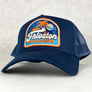 Może przedstawiać: Czapka trucker w kolorze granatowym z naszywką przedstawiającą palmę i napis "Thloston Paradise" w kolorze pomarańczowym i białym.