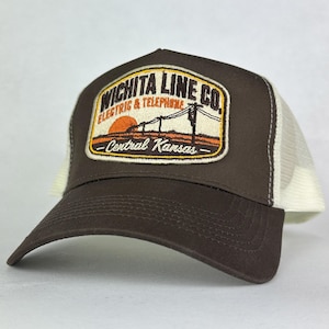 May include: A brown and off-white trucker hat featuring an embroidered patch on the front. The patch displays a sunset landscape with utility poles and the text WICHITA LINE CO. ELECTRIC & TELEPHONE CENTRAL KANSAS in black and orange lettering.