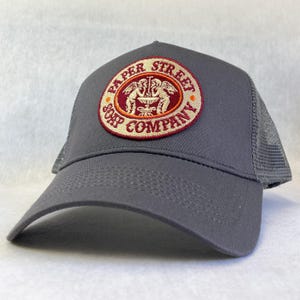 Puede incluir: Una gorra de camionero gris con un parche bordado en burdeos y dorado que dice "Paper Street Soap Company".