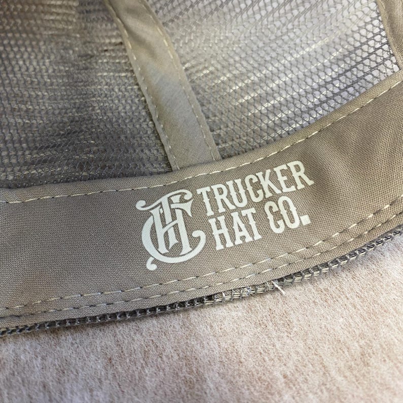 K&ouml;nnte beinhalten: Nahaufnahme des Inneren einer Trucker-M&uuml;tze. Die M&uuml;tze hat eine Netzr&uuml;ckseite und ein beiges Schwei&szlig;band mit dem wei&szlig;en Text "TRUCKER HAT CO.". Das Design der M&uuml;tze beinhaltet ein dekoratives "H"-Logo.