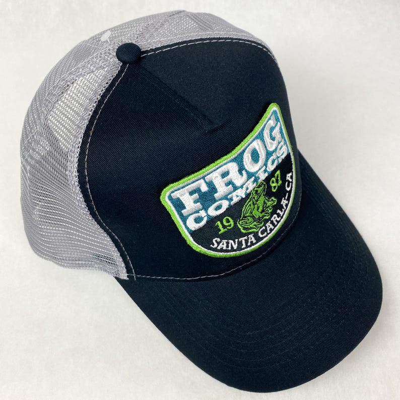 May include: Black and grey trucker hat with a green, white, and blue embroidered patch that says "Frog Comics 1987 Santa Carla CA" and a green frog illustration.