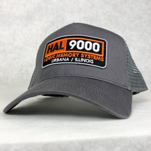 Peut inclure: Un chapeau de camionneur gris et noir avec un patch orange et blanc qui indique "HAL 9000 LOGIC MEMORY SYSTEMS URBANA / ILLINOIS"