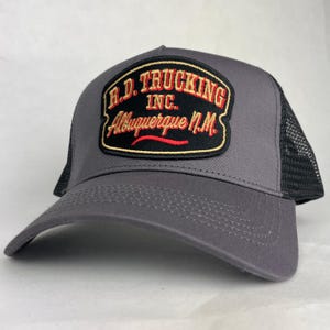 May include: A grey trucker cap featuring a black patch on the front with embroidered red and gold text that reads R.D. TRUCKING INC. Albuquerque N.M. The cap has a solid grey front panel and a black mesh back panel with a curved brim.