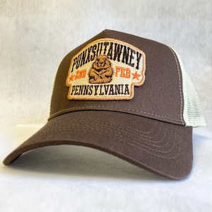 May include: Brown and white trucker hat with a patch that says "Punxsutawney, PA, 2nd Feb" and features a groundhog.