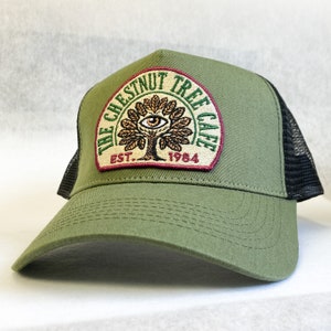 Op de afbeelding: Een groene truckercap met een zwart mesh achterpaneel. De pet heeft een patch met een boom met een oog in het midden en de tekst "The Chestnut Tree Cafe Est. 1984".