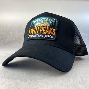 Op de afbeelding: Zwarte truckercap met een blauw en geel geborduurd patch met de tekst "Welkom in Twin Peaks Bevolking 51201".