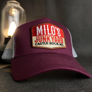 Könnte beinhalten: Burgunderfarbene Trucker-Kappe mit weißem Netzrücken. Die Kappe hat einen roten, weißen und braunen Aufnäher mit dem Text "Milo's Junkyard Castle Rock, ME."