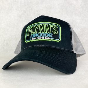 Könnte beinhalten: Schwarze Trucker-Cap mit gebogenem Schirm und Mesh-Rückseite. Auf der Vorderseite steht "FLYNN'S ARCADE" in Neon-Grün und Blau. Der Text "9543 CULVER BLVD" befindet sich darunter.
