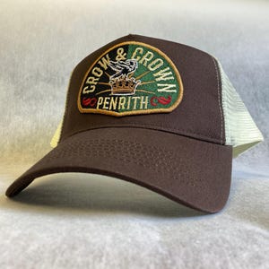 Puede incluir: Gorra de camionero marrón y blanca con un parche bordado verde y dorado que dice "Grow & Crown Penrith".