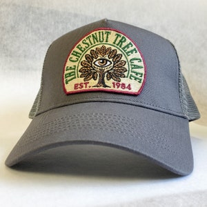 May include: Grey trucker hat with a patch featuring a stylised tree with an eye, the text "The Chestnut Tree Cafe", and the text "Est. 1984".