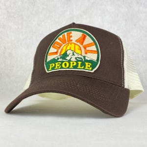 Peut inclure: Casquette de camionneur marron et blanche avec un patch brodé vert, orange et jaune qui dit "Love All The People" avec un graphique de montagne et de soleil.