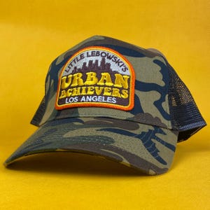 Könnte beinhalten: Tarnfarbene Trucker-Mütze mit gebogenem Schirm und Mesh-Rückseite. Der vordere Aufnäher zeigt den Text "LITTLE LEBOWSKI'S URBAN ACHIEVERS LOS ANGELES" in gelben und weißen Buchstaben mit einem orangefarbenen Rand. Die Mütze ist vor gelbem Hintergrund platziert.