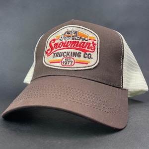 Op de afbeelding: Een bruine truckerpet met een witte mesh achterkant en een centrale geborduurde patch. De patch toont een oranje vrachtwagengrafiek boven de rode tekst "Snowman's TRUCKING CO." en "EST. 1977" in een rood ovaal, op een crèmekleurige achtergrond.