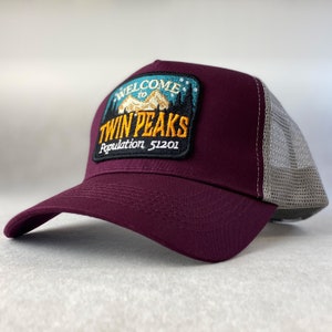 May include: Burgundy trucker hat with a black and yellow embroidered patch that says "Welcome to Twin Peaks Population 51201". The patch features a mountain range and a cabin.