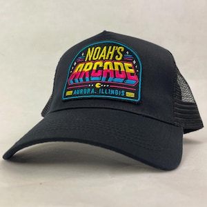 May include: A black trucker cap featuring an embroidered patch on the front. The patch displays the text NOAH'S ARCADE and AURORA, ILLINOIS in yellow, pink, and blue lettering with a small yellow crescent moon icon on a black background.