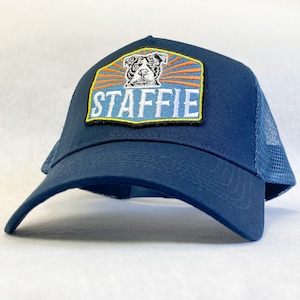 Op de afbeelding: Een marineblauwe truckerpet met een geborduurde patch aan de voorzijde. De patch toont een zwart-witte illustratie van een hondenkop boven het woord STAFFIE in witte letters, tegen een achtergrond van oranje en blauwe zonnestralen.