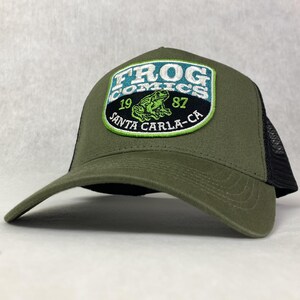 May include: A green trucker hat with a black mesh back. The hat has a large embroidered patch on the front that says "Frog Comics" with a green frog and the text "19 Santa Carla-CA 87".