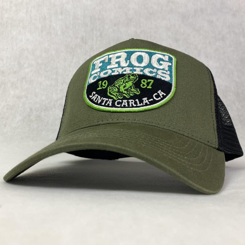 May include: A green trucker hat with a black mesh back. The hat has a large embroidered patch on the front that says "Frog Comics" with a green frog and the text "19 Santa Carla-CA 87".
