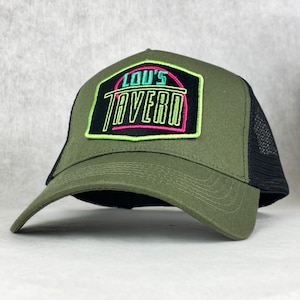 Puede incluir: Gorra de camionero verde oliva con parte trasera de malla negra y visera curva. El parche frontal muestra el texto "LOU'S TAVERN" en verde neón y rosa, delineado en negro. Un accesorio de moda.