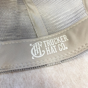 May include: Close-up of the back of a gray trucker hat with a white embroidered logo that says "Trucker Hat Co."