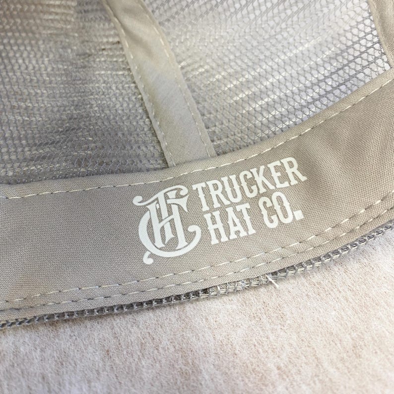 May include: Close-up of the back of a gray trucker hat with a white embroidered logo that says "Trucker Hat Co."