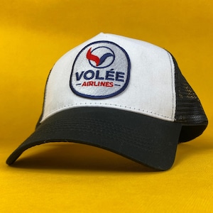 May include: A trucker hat with a white front panel and a black mesh back. The hat features a patch with the text "VOLÉE AIRLINES" in a blue circle with a red and blue design. The brim is black.