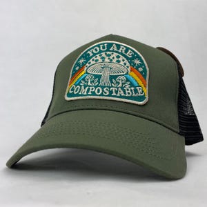 May include: An olive green trucker cap with a black mesh back and a central embroidered patch. The patch features a mushroom illustration, a rainbow, and the text "YOU ARE COMPOSTABLE" in white lettering against a dark teal background.