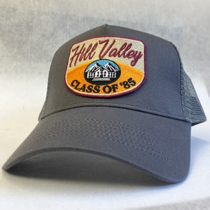 Pode incluir: Boné de caminhoneiro cinza com um patch bordô, laranja e branco que diz "Hill Valley Class of '85".