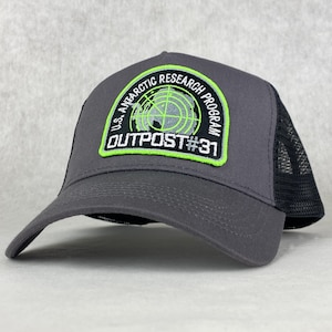 Op de afbeelding: Een grijze en zwarte truckercap met een groen en wit geborduurd patch met de tekst "U.S. Antarctic Research Program Outpost #31".