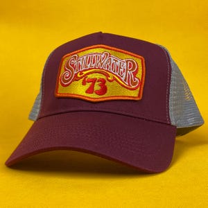 May include: A burgundy trucker cap with a grey mesh back and a curved peak. The front panel displays a rectangular patch with the word "Stillwater" and the number "73" in red and gold.