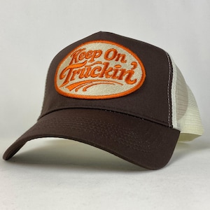 May include: A brown trucker hat with a white mesh back. The front of the hat features an oval patch with the words "Keep On Truckin'" in orange script.