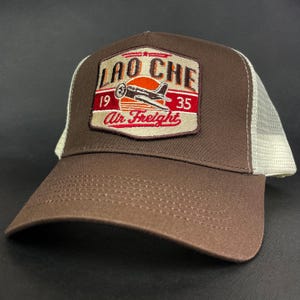 May include: A brown trucker hat with a white mesh back and a front patch featuring an embroidered airplane graphic, a red sun, and the text Lao Che 1935 Air Freight. The hat has a curved brim with visible stitching and a structured front panel.