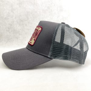 K&ouml;nnte beinhalten: Graue Trucker-Cap mit Mesh-R&uuml;ckseite und gebogenem Schirm. Auf der Vorderseite befindet sich ein rechteckiger Aufn&auml;her mit dem Wort "Ranch" in bordeauxfarbenen Buchstaben. Ein braunes Etikett ist an der R&uuml;ckseite befestigt.